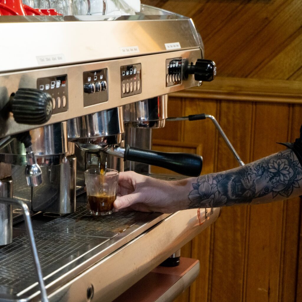 Steph's tattooed arm holding espresso glass under coffee stream
