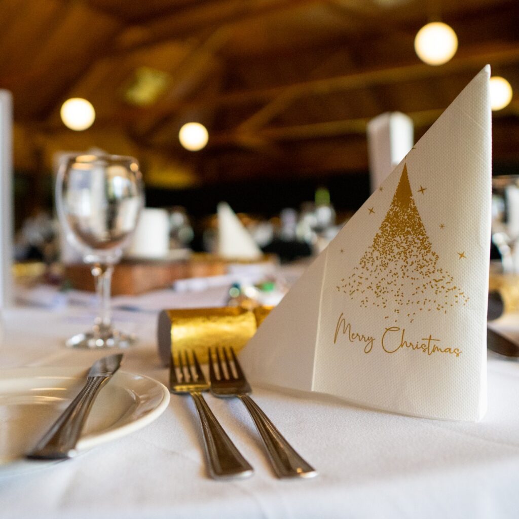 Table set with cutlery, side plates, wine glasses and festive decorations with napkin with 'Merry Christmas' and tree printed in gold as primary focus