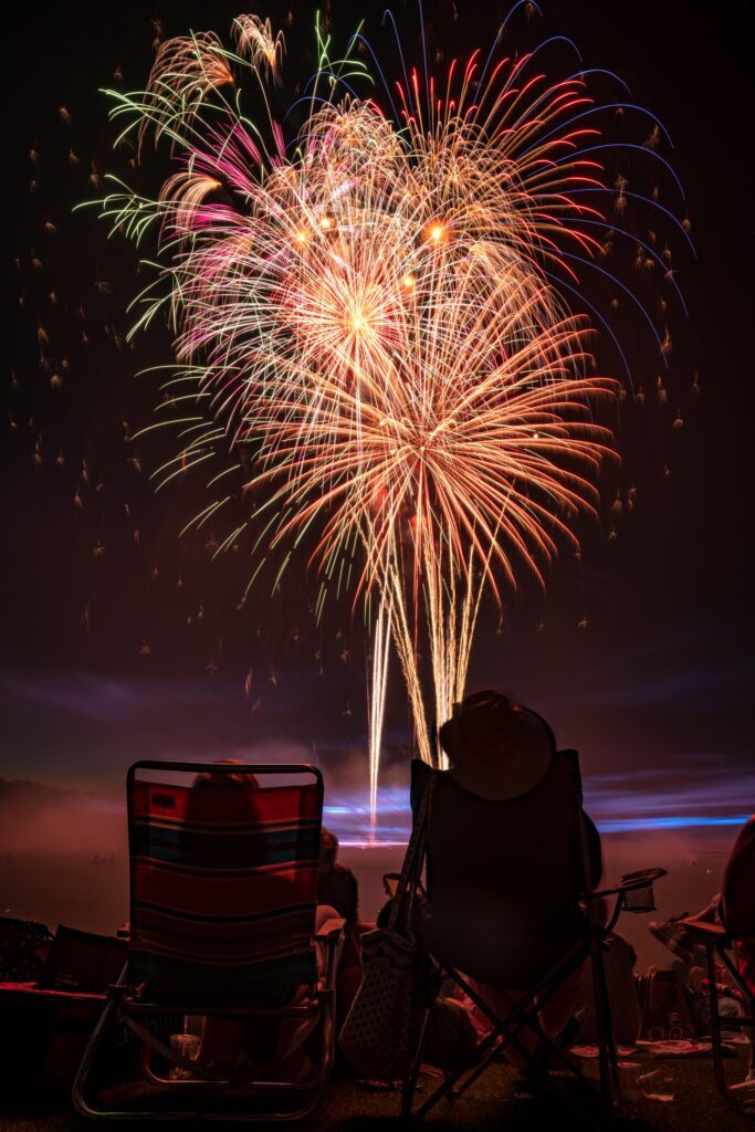 People sitting in beach chairs looking up at a burst of fireworks