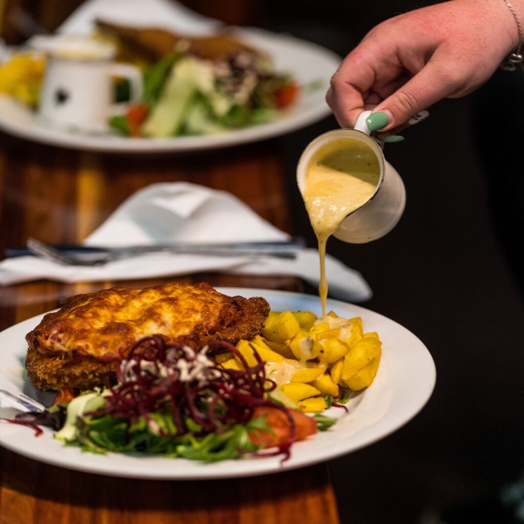 Woman pouring creamy garlic sauce on hot chips beside chicken parmi