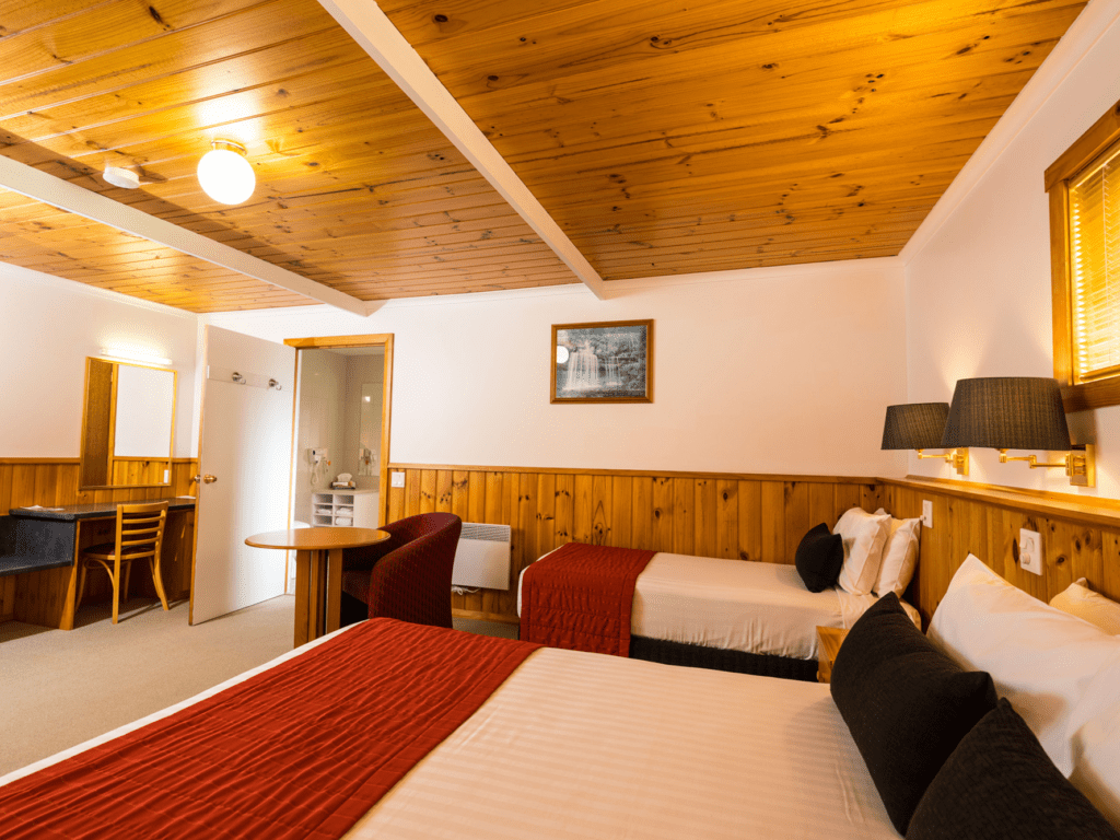 Wood panelled standard Disability Access room with queen and single bed, moody lighting and small table with tub chair. Bathroom entry is visible and large mirror over working space with standard chair next to luggage storage